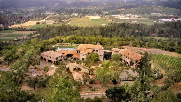 Aerial view of a large Mediterranean-style estate set within landscaped grounds and surrounded by vineyards, orchards, farmland, and wooded hills. The complex consists of several low-rise buildings with terracotta tiled roofs arranged around courtyards and garden spaces, with pale stucco or stone-toned walls and mature trees.