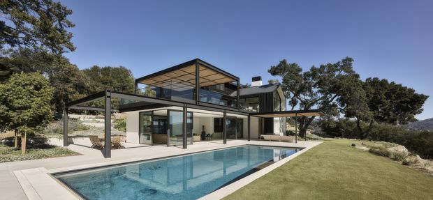 Contemporary multi-level residential house with extensive floor-to-ceiling glazing, dark slim structural framing, broad cantilevered roof planes, and a large rectangular swimming pool. The home sits within a landscaped, slightly sloping site with mature trees and lawn, and has strong indoor-outdoor connections through covered terraces and open living areas.