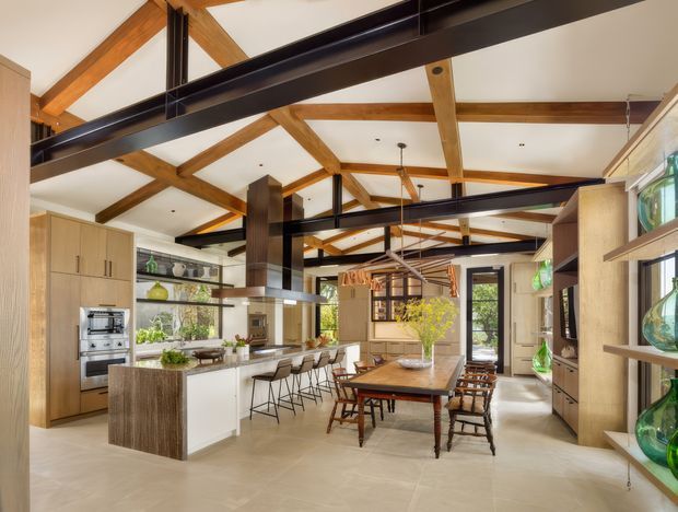 A bright, expansive open-plan kitchen and dining area with a large central island, integrated appliances, timber cabinetry, and a long dining table. The room features exposed warm timber roof trusses, black steel beams, clerestory-like glazing, pendant lighting, and display shelving with glass vessels. Large windows and doors provide garden views, while pale flooring and walls create an airy contemporary interior.