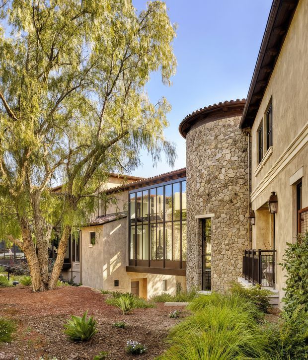 Exterior view of a warm, low-rise architectural residence arranged around a landscaped courtyard. The building combines pale stucco walls, a prominent rounded stone-clad tower, dark timber roof detailing, and a tall glazed connector with dark-framed windows. Drought-tolerant planting, mature willow trees, and a clear blue sky contribute to a Mediterranean-inspired, tranquil setting.