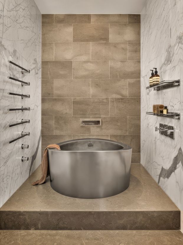 Luxury contemporary bathroom featuring a freestanding brushed-metal bathtub set on a raised platform within a tiled wet-room enclosure. The rear wall uses large warm taupe stone-look tiles in a staggered layout, while the side walls are clad in dramatic white marble-look slabs with grey veining. Multiple dark metal shower outlets line the left wall, with open metal shelves and bath products on the right. The overall composition is sculptural, refined, and spa-like.