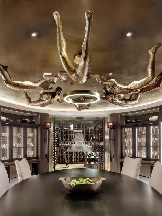 Luxurious private dining and wine room with a large dark round dining table, upholstered dining chairs, illuminated glass-front display cabinetry, and a glass-enclosed wine cellar at the rear. The room features a dramatic sculptural antler-like chandelier, a luminous circular pendant, dark timber joinery, metallic wall finishes, and warm recessed lighting.