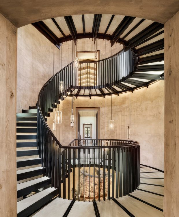 A dramatic multi-level spiral staircase and landing viewed through a timber-framed opening. The stair has dark metal balustrades and treads, pale stone or concrete-like surfaces, exposed dark structural beams, hanging pendant lights, and textured warm neutral walls. The composition emphasizes a tall vertical circulation void with curved forms and layered landings.