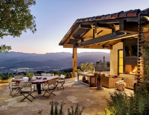 A rustic, timber-framed outdoor terrace overlooking a broad mountainous valley. The covered entertaining area has exposed timber rafters and beams, a tiled pitched roof, stone-paved flooring, an outdoor dining table with chairs, and a lounge area arranged around a built-in fireplace or hearth. Warm evening light highlights the natural materials and expansive elevated outlook.