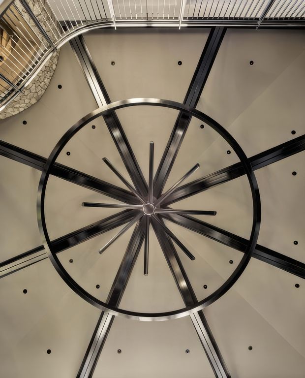 Upward-looking view of a dramatic circular architectural ceiling feature, with a large dark metal ring and radial beams or ribs converging at a central hub. The surrounding ceiling is a warm off-white plastered surface with recessed spotlights, while a curved metal balustrade and exposed stone wall are visible at the upper edge, suggesting a multi-level interior atrium or stairwell. The composition is highly geometric and symmetrical.