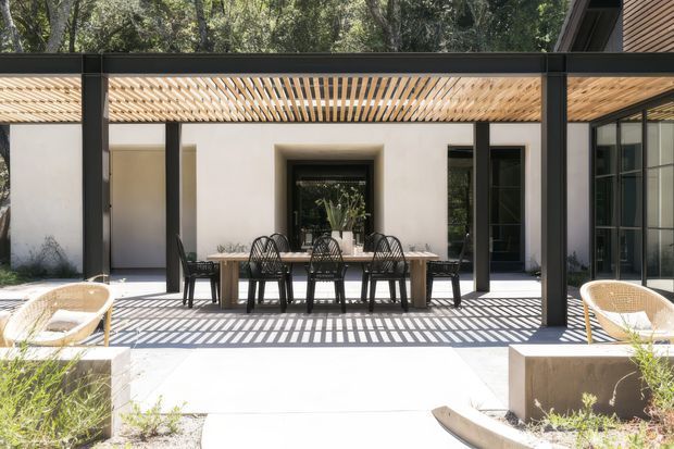 A contemporary courtyard dining area viewed from an outdoor patio. A long timber dining table with black woven chairs sits beneath a timber slatted pergola supported by dark structural posts. White rendered walls, large dark-framed glazed openings, and surrounding mature greenery create a bright, geometric indoor-outdoor setting. Strong linear shadows from the pergola fall across the pale paving, with built-in planter edges and woven lounge chairs in the foreground.