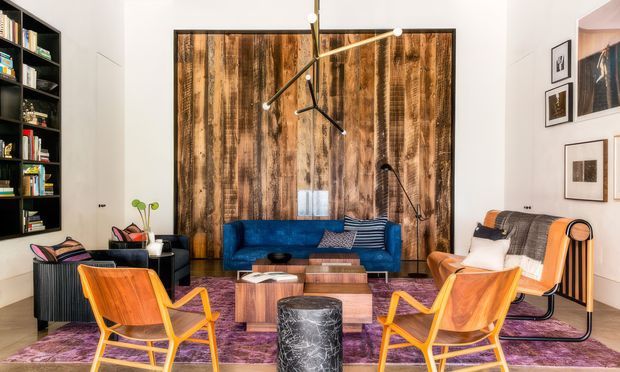 A stylish contemporary living room with a vivid blue sofa, two orange timber lounge chairs, a black-and-white lounge chair, a low timber coffee table, and a cylindrical dark stone or marble side table. The room features a large rustic reclaimed-timber feature wall, extensive black built-in shelving, white walls, framed artwork, a purple area rug, and a sculptural modern pendant light. The overall composition is eclectic with strong mid-century-inspired furniture and warm timber accents.