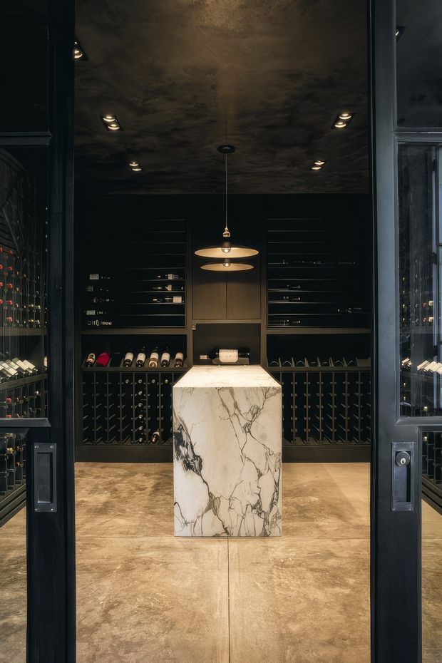 A dark, sophisticated wine room viewed through open black-framed doors. Full-height black wine racks and bottle storage line the walls, while a sculptural white marble island with dramatic grey veining anchors the centre. The ceiling has a mottled dark finish with recessed spotlights and a pair of warm pendant lights. The floor is finished in large warm taupe-brown slabs, creating a moody, luxurious cellar atmosphere.