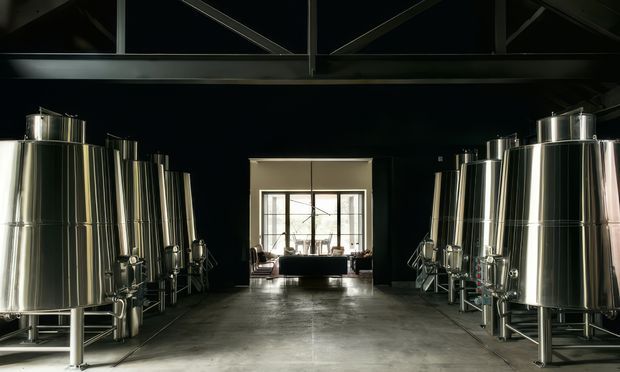 Interior view of a large industrial brewing or fermentation facility, with rows of polished stainless-steel tanks lining both sides of a wide concrete-floored space. A dark exposed structural roof and clerestory-like glazing frame the upper portion, while a bright glazed opening and small seating area appear at the far end.
