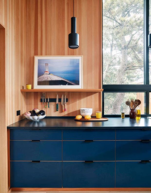 A richly detailed kitchen vignette with deep navy-blue base cabinetry and a dark countertop set against vertical warm timber wall panelling. A floating timber shelf, framed artwork, hanging black pendant light, large black-framed window, knife rack, utensils, bowls and small countertop items add character. The space feels compact, warm and thoughtfully designed, with views of trees through the window.