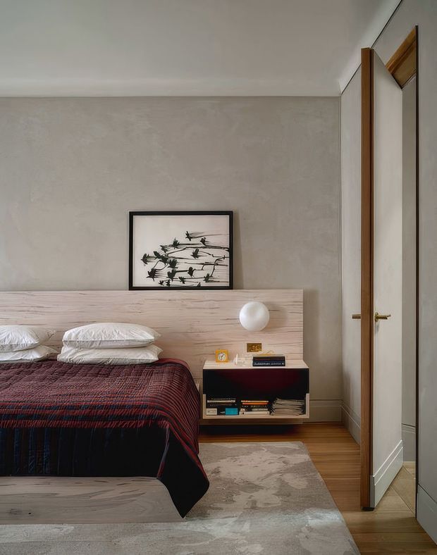A warm, refined bedroom with a low pale timber bedhead, layered white pillows, and a dark red and navy patterned bedcover. A black open bedside cabinet holds books and small objects, while a spherical white lamp sits on the timber headboard. The room features softly textured greige plaster walls, timber flooring, a muted patterned rug, framed botanical artwork, and full-height mirrored wardrobe doors with timber frames.