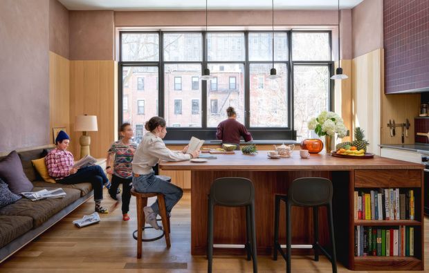 Warm, eclectic open-plan kitchen and living space with a large timber island and breakfast bar, two stools, integrated shelving, dark cabinetry, pendant lights, and a broad industrial-style window wall. A sofa and several people occupy the living area, while the kitchen includes a tiled backsplash, countertop appliances, and open shelving. The room has timber flooring, soft pink-beige walls, and a relaxed, social atmosphere.