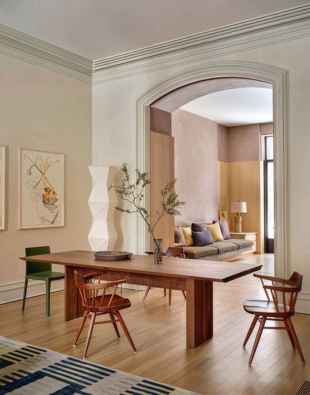 Elegant open-plan dining area with a large mid-century timber dining table and matching wooden chairs, set against warm pale walls and timber flooring. A sculptural white floor lamp, framed artwork, vase with branches, and patterned rug add visual interest. Through a wide, heavily moulded arched opening is a refined sitting room with built-in timber seating, cushions, and a tall window. The space combines traditional architectural detailing with curated mid-century furnishings and warm contemporary styling.