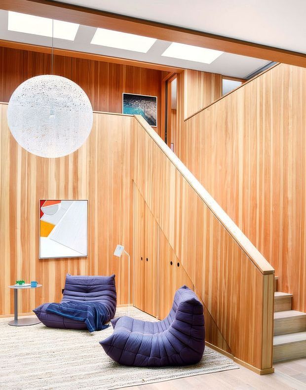 A warm, wood-panelled interior centred on a staircase with integrated vertical timber storage cabinetry. Two deep purple lounge chairs sit on a pale textured rug beside a small round side table. A large spherical pendant light, skylights, framed artwork, and a diagonal stair balustrade create a sculptural, mid-century-inspired living area.