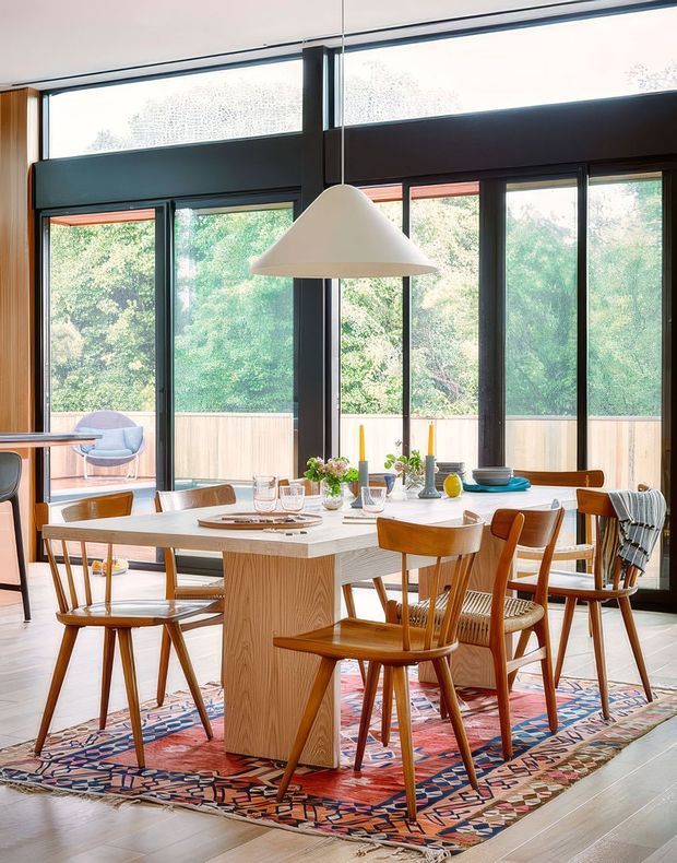 Bright dining area with a large rectangular timber dining table surrounded by mid-century style wooden chairs, set on a colourful patterned rug. The space features expansive dark-framed glazing and sliding doors opening to a leafy outdoor setting, timber wall panelling, a contemporary white pendant light, and simple table styling.