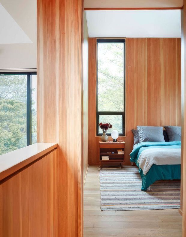 A compact bedroom with extensive vertical timber wall panelling and cabinetry, large black-framed windows overlooking trees, and a neatly made bed with teal bedding. A small timber bedside table, vase, books, and a striped area rug add warmth. The room has a bright, calm atmosphere and a strong connection to the surrounding greenery.