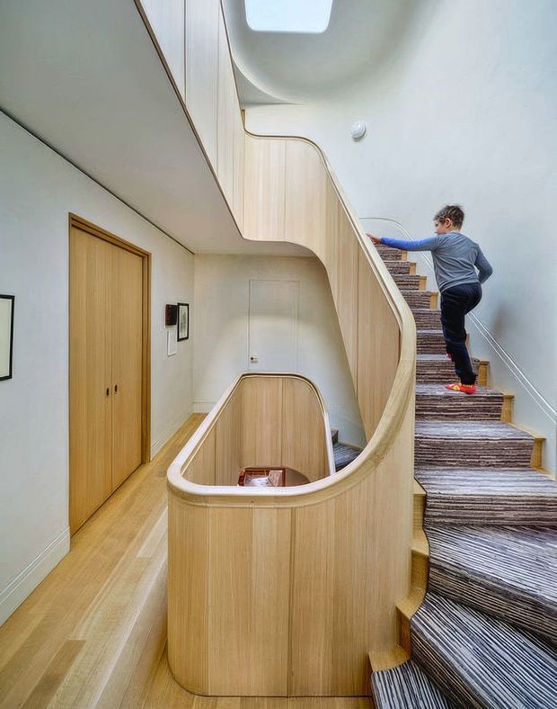 Interior view of a sculptural curved staircase and stairwell, with pale timber panelling and matching curved balustrades wrapping around a central opening. The stairs are carpeted in a striped grey-brown textile, with pale timber flooring and doors along the adjacent hallway. A child is ascending the stairs, and a roof skylight brings natural light into the tall stairwell.