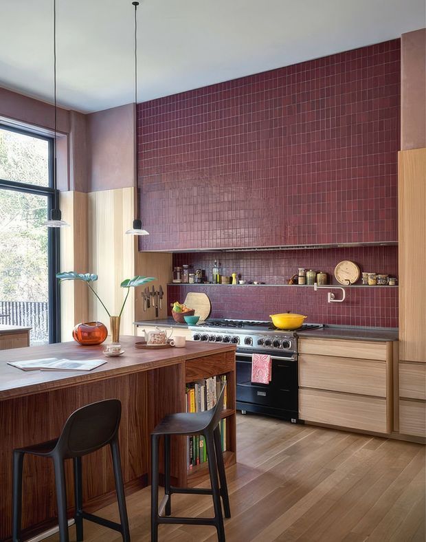 A warm, design-focused kitchen with a tall burgundy-red tiled backsplash and matching upper wall, light timber cabinetry, a dark timber island with integrated open shelving, and a freestanding stainless-steel range cooker. Black bar stools, pendant lights, a large window, and curated countertop objects complete the mid-century-influenced interior.