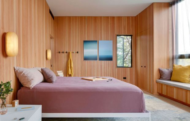 Warm, wood-panelled bedroom with a low upholstered bed dressed in muted mauve bedding, built-in timber wardrobes and a window seat with cushions. Vertical timber wall panelling, integrated wall lights, small framed artwork, and a large window create a refined, cozy interior with natural light.