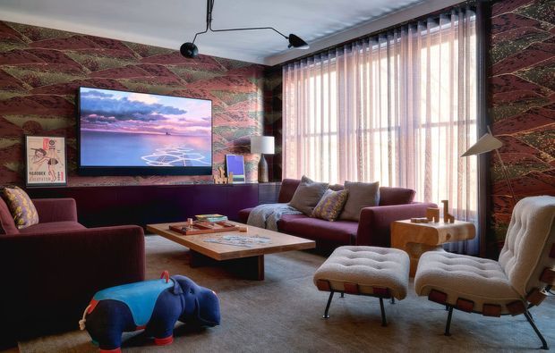A richly layered mid-century-inspired living room with burgundy upholstered seating, patterned feature walls, a low timber coffee table, sculptural lounge chairs, ottomans, and a large television. Full-height vertical blinds filter daylight through the expansive window, while an unusual pendant light and colourful artwork add playful character.