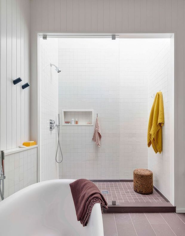 Bright contemporary bathroom featuring a large open walk-in shower with white tiled walls, a dark mauve tiled shower floor, recessed shelf, handheld shower, and glass screen. A freestanding white bathtub occupies the foreground with a draped mauve throw. Vertical pale wall panelling, dark fixtures, and mustard-yellow towels add contrast, while a small upholstered stool sits on the raised shower platform.
