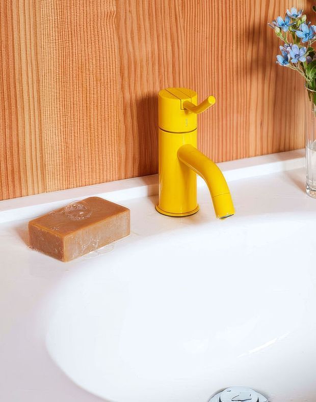 Close-up view of a bright white bathroom basin with a vivid yellow single-lever faucet. The basin is set against a vertical warm timber feature wall, with a translucent amber soap bar on the countertop and a small glass vase of blue flowers at the side. The image is bright, playful, and colour-focused, with the faucet as the main product feature.