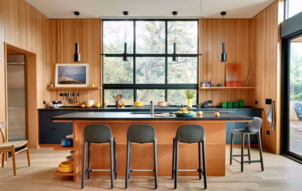 Contemporary kitchen with a large central island and seating for three, surrounded by warm vertical timber wall panelling. The space features dark cabinetry, a dark countertop, integrated stainless-steel refrigerator, open shelving, pendant lights, and expansive black-framed glazing overlooking trees. A dining or outdoor area is visible through the glazed door on the right.