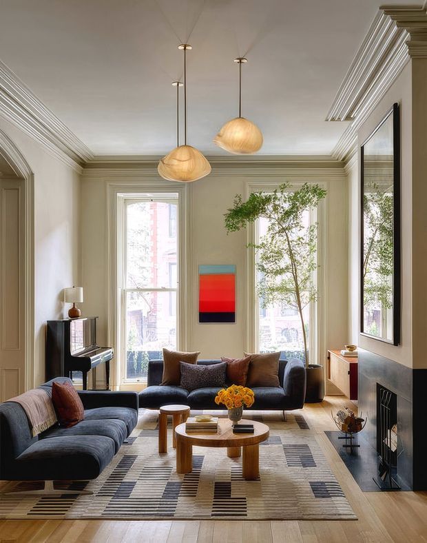 A sophisticated, generously proportioned living room with tall windows, ornate wall and ceiling mouldings, and a neutral taupe palette. Two sculptural pendant lights hang over a seating arrangement of dark blue-grey sofas and leather accent cushions. A grand piano, colourful abstract artwork, patterned area rug, round timber coffee table, fireplace, and leafy indoor branches create a refined, layered interior.