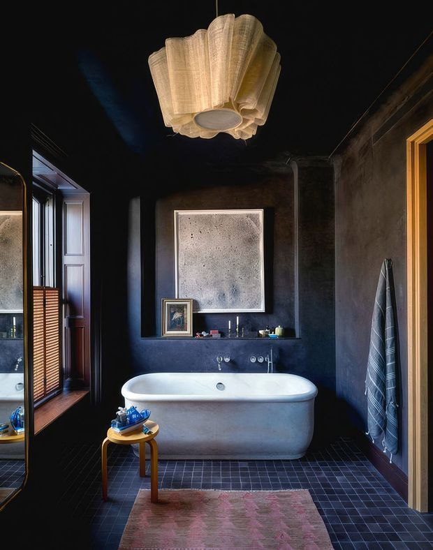 A moody, dark-toned bathroom with a freestanding white bathtub, black tiled floor, dark plaster walls, timber-framed windows and door, and a sculptural fabric pendant light. A towel hangs beside the tub, with a small timber stool and bath accessories in the foreground. The room combines dramatic lighting with rustic, textured finishes and warm timber accents.