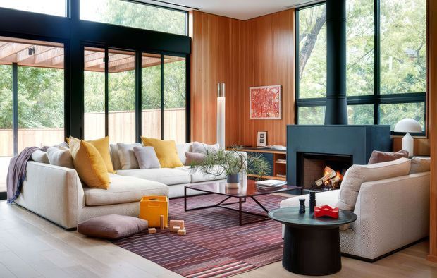A warm, contemporary living room with a large pale sectional sofa, armchair, low coffee table, and striped area rug arranged around a freestanding black wood-burning fireplace. Full-height black-framed glazing and a glazed corner open to a leafy outdoor setting, while vertical timber wall panelling adds warmth. The room is bright, comfortable, and connected to a covered outdoor terrace.