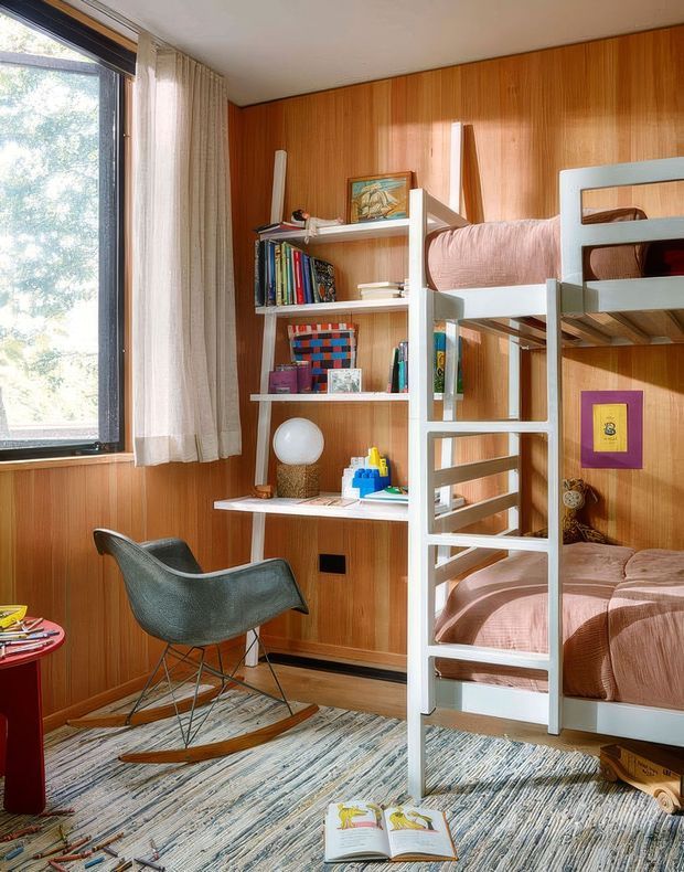 A compact, warmly lit children's bedroom with built-in bunk beds, a white ladder, open shelving filled with books and toys, and a small desk beneath a large window. The room features extensive vertical timber wall panelling, pale bedding, a dark rocking chair, and a textured patterned rug. Natural light enters from the window, creating a cozy, playful reading and sleeping space.