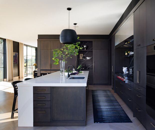 A sophisticated open-plan kitchen with a large white stone island, dark timber cabinetry, integrated appliances, open shelving, and pendant lighting. The kitchen opens toward a bright living or dining area with expansive glazing and curtains. A striped dark runner, greenery, and restrained furnishings add contrast to the refined scheme.