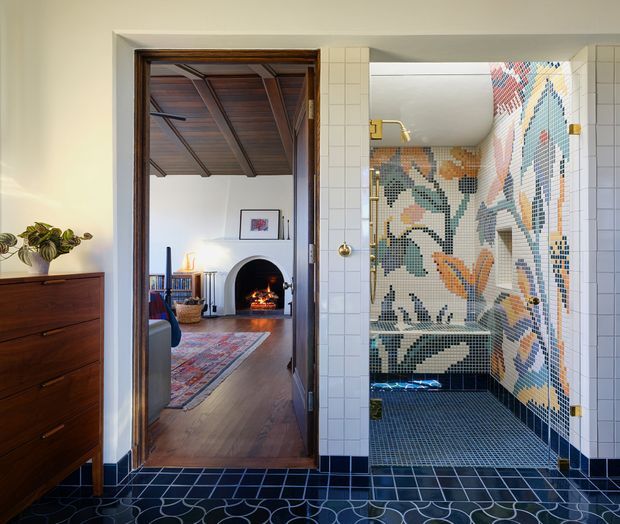 A richly detailed bathroom viewed from a tiled entry area, featuring a large colourful mosaic wall depicting abstract botanical and geometric forms, dark blue tiled flooring with curved line motifs, and white square wall tiles. Through the open timber-framed doorway, a warmly furnished living room with timber flooring, a fireplace, rug and mid-century-style furniture is visible. The bathroom includes brass-toned hardware and a glass shower enclosure.