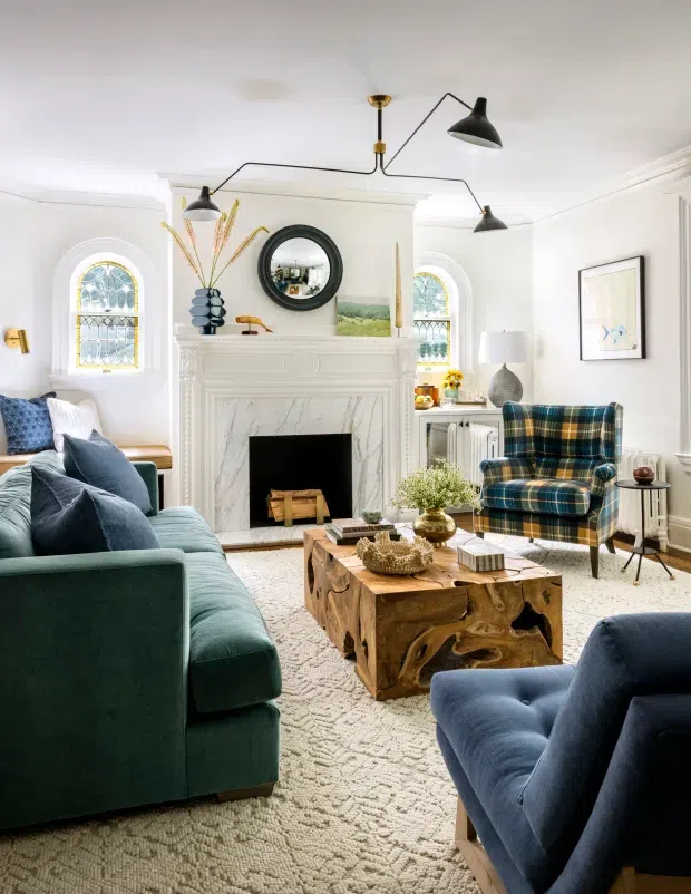 A bright, traditionally detailed living room with a white marble fireplace surround, ornate mantel, arched leaded-glass windows, decorative wall mouldings and a round mirror. Deep teal sofas and a blue-green plaid armchair surround a sculptural natural timber coffee table, with a textured neutral rug and contemporary black multi-arm pendant lighting.