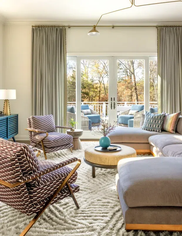 Bright, layered living room with a large pale sectional sofa, two patterned mid-century-style lounge chairs, a round upholstered coffee table, textured cream rug, blue sideboard, and floor-to-ceiling curtains. White-framed glazed doors open to a balcony or terrace with additional seating and leafy trees beyond. A sculptural brass ceiling light and soft neutral palette create an inviting, refined atmosphere.