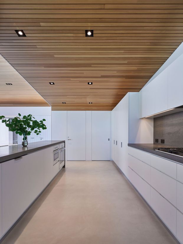 A sleek, contemporary galley kitchen with extensive white handleless cabinetry on both sides, dark stone-look benchtops, integrated appliances, and a warm slatted timber ceiling with recessed spotlights. The open aisle leads toward tall white storage and a concealed door, while a potted plant adds a small natural accent.