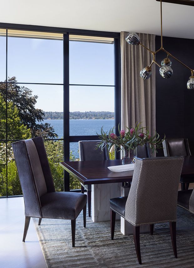 A sophisticated dining room with a dark timber dining table, upholstered dining chairs, a sculptural branching pendant light, and a large patterned area rug. Full-height black-framed glazing overlooks a blue body of water and wooded shoreline. Neutral finishes, dark furniture, and floral greenery create a refined, dramatic setting.