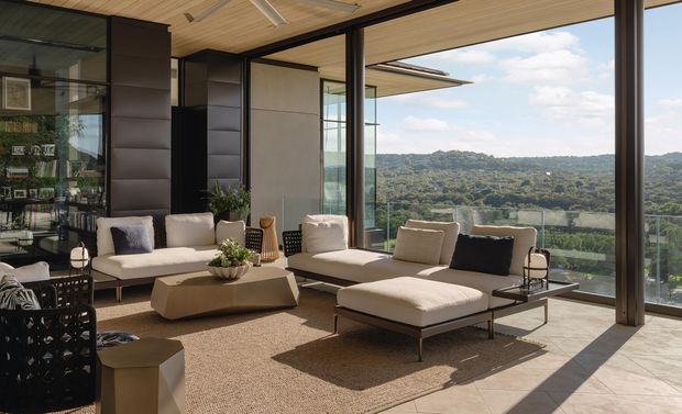 A bright, contemporary living room with expansive floor-to-ceiling glazing overlooking a wooded landscape. The space features a large neutral sectional-style seating arrangement with chaise, a low sculptural coffee table, textured area rug, dark framed glazing, dark built-in cabinetry, and a warm timber-lined ceiling. The open indoor-outdoor feel and abundant natural light create a refined, airy lounge environment.