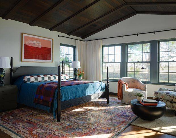A warmly lit bedroom with a dark wood-paneled pitched ceiling, large multi-pane windows, and a four-poster bed dressed in blue bedding and a patterned throw. Layered Persian-style rugs cover the floor, while a curved upholstered armchair, round black coffee table, bedside furniture, artwork, and a floor lamp create a richly styled, eclectic interior.