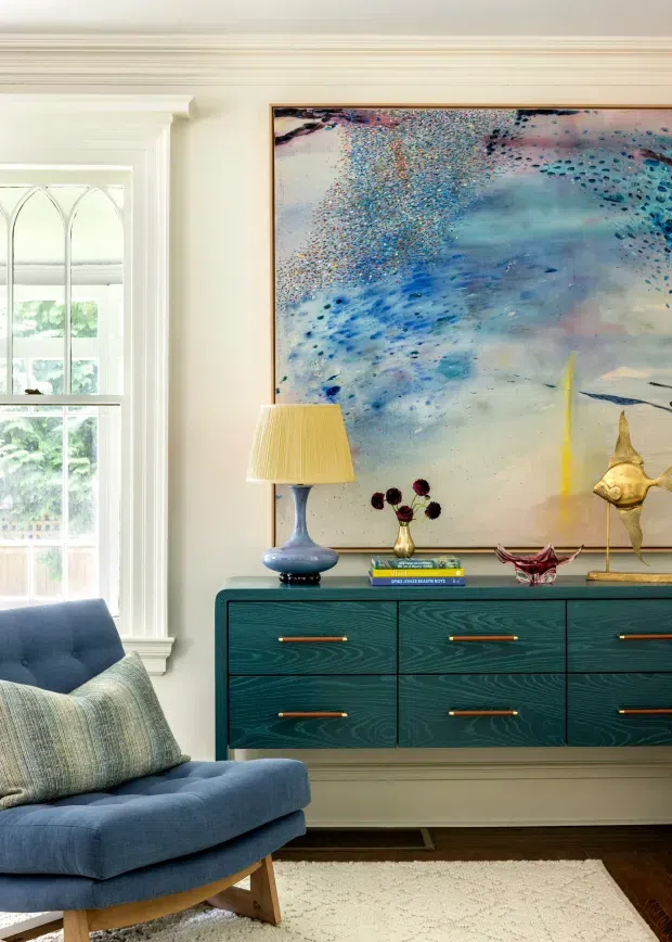 A refined living room featuring a deep teal built-in or freestanding six-drawer sideboard, a blue upholstered lounge chair with textured cushions, and a large abstract blue artwork. The room has traditional architectural detailing with ornate layered cornice mouldings, white trim, and a tall divided window. Brass-toned drawer pulls, a sculptural lamp, decorative objects, and a light neutral rug add contrast to the rich blue-green furnishings and dark timber floor.