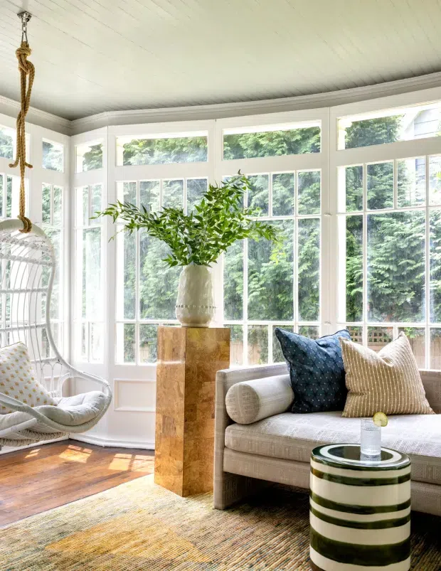 Bright enclosed sunroom with extensive white-framed multi-pane glazing, a painted plank ceiling, warm timber flooring, and a neutral upholstered sofa. A hanging wicker chair, patterned cushions, striped cylindrical side table, woven rug, and a tall timber pedestal with a ceramic vase add relaxed contemporary-coastal character. Lush greenery is visible outside the windows.