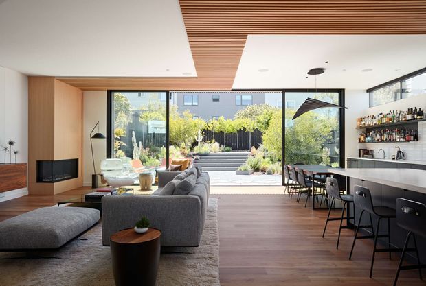 Open-plan contemporary living and entertaining space with a grey sectional sofa facing a wall-mounted fireplace and television, adjoining a long kitchen island and home bar. Full-height glazed doors open to a planted courtyard, while warm timber ceiling slats and timber cabinetry contrast with white walls, dark window frames, and dark wood flooring.