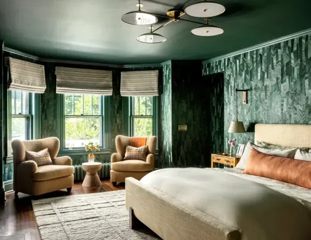 A richly styled bedroom with deep green botanical-patterned wallpaper and matching ceiling, three tall windows with woven Roman blinds, a large upholstered bed, two plush armchairs, a small side table, and layered neutral and rust-toned textiles. Contemporary decorative pendant lighting and warm natural light create a refined, cozy atmosphere.
