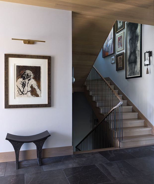 A refined interior stairway and landing with a sculptural console table, framed artwork, a dark slate floor, and a timber-clad sloping ceiling. The staircase features light timber treads, dark vertical metal balustrades, and a curved handrail, while gallery-style artwork and wall-mounted lighting add a sophisticated decorative layer.