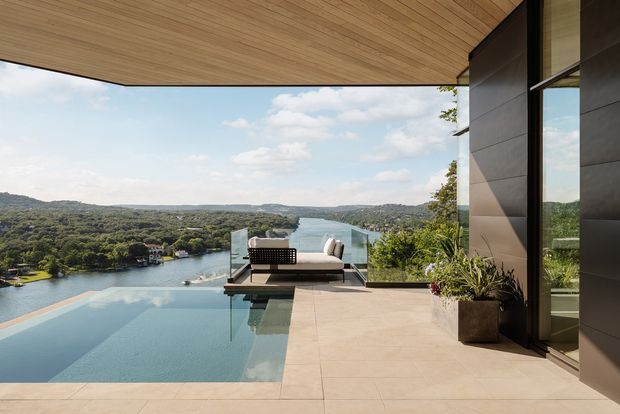 A contemporary waterfront terrace overlooking a broad river or estuary, with an infinity-edge swimming pool in the foreground, outdoor lounge seating, pale tiled paving, dark exterior wall cladding, and a warm timber-lined soffit. Large glazing reflects the landscape, while landscaped hills and water extend into the distance.