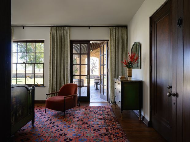 A refined traditional entry hall or living area with dark timber flooring, a richly patterned red and blue rug, a sculptural rust-red armchair, and a dark wood console cabinet. Full-height patterned curtains frame black-grid windows and glazed double doors opening to an outdoor terrace. A large dark timber paneled door dominates the right side, while natural light creates a warm, elegant atmosphere.