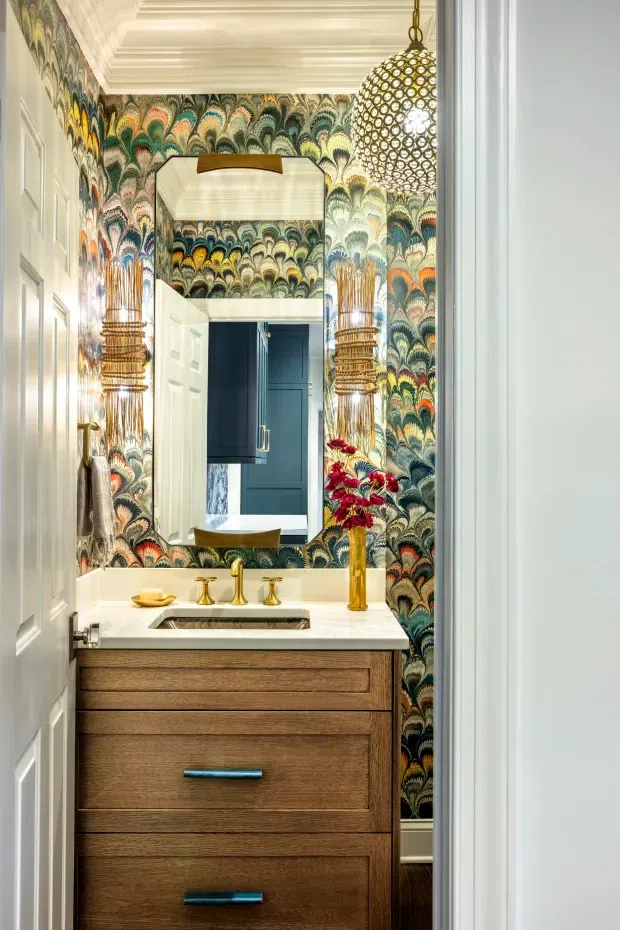 A compact, richly decorated powder room viewed through an open doorway. The room features bold multicoloured patterned wallpaper, a warm timber vanity with brushed blue-green pulls, a white stone benchtop and integrated basin, brass tapware, a large brass-framed mirror, and an ornate spherical pendant light. A dark blue room is visible in the mirror and doorway beyond.