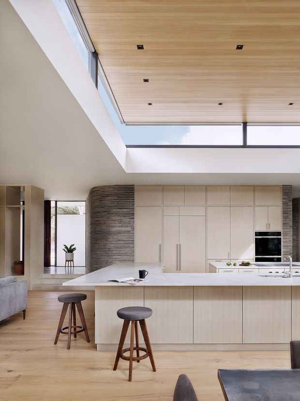A bright contemporary kitchen viewed from the adjoining living area. The room features a large white island with seating for two, pale timber floor-to-ceiling cabinetry, integrated black appliances, a white benchtop, a curved textured brick feature wall, and a timber-lined ceiling with recessed spotlights. A large roof opening or clerestory brings in daylight and creates a strong connection to the sky.