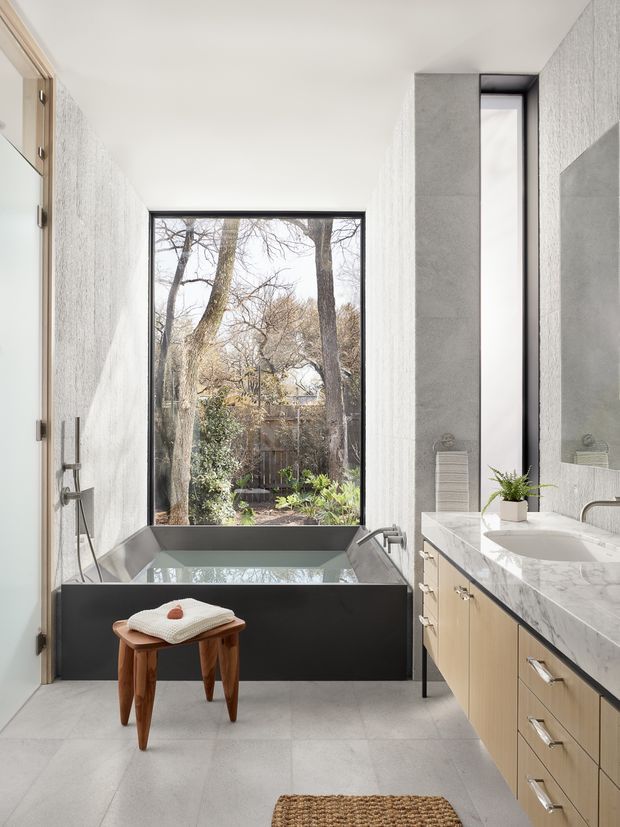 Bright contemporary bathroom with a freestanding dark bathtub positioned in front of a large black-framed window overlooking trees. The room features pale textured wall surfaces, large light-grey floor tiles, a marble-look vanity benchtop with light timber cabinetry, brushed metal tapware, a tall narrow window, and a small wooden stool.
