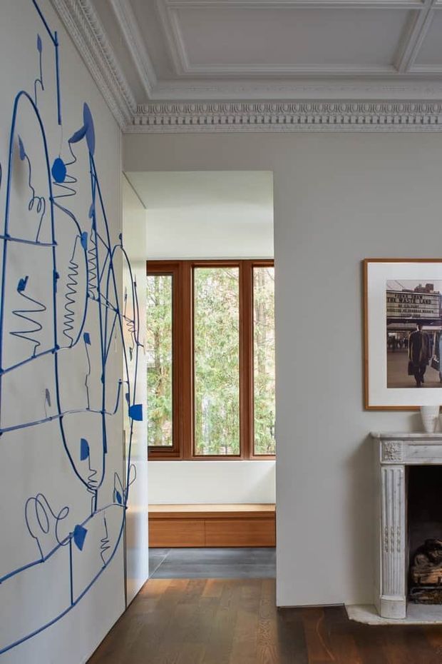 A refined residential interior hallway or living area with ornate white decorative ceiling cornices and mouldings, pale grey walls, dark timber flooring, and a classic white fireplace surround. A large blue abstract mural occupies the left wall, while a framed monochrome artwork hangs above the fireplace. Through the opening, a bright window seat or built-in timber bench is visible, with timber-framed glazing overlooking trees.